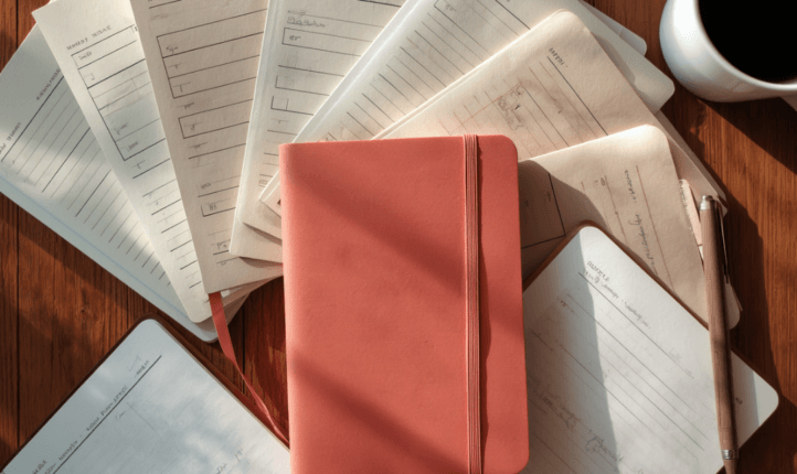 Flat lay of scattered paperwork and notebooks on a wooden desk, with a coral journal and a cup of coffee — representing the juggling act of supporting multiple executives.